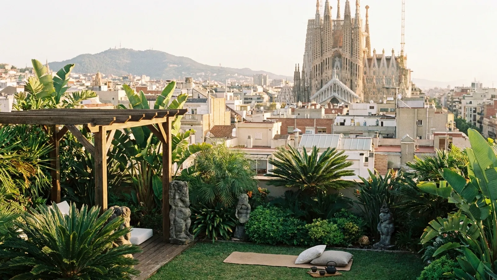 Serene view of Barcelona rooftops and a distant mountain range at sunrise, with a quiet, leafy terrace in the foreground.